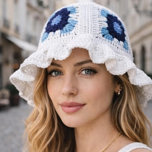 以下が含まれることがあります： 白いかぎ針編みのバケットハット、フリル付きのつば。帽子は青色の濃淡の花柄です。この手作りの帽子は、日焼け防止のためのスタイリッシュなアクセサリーで、どんな服装にも手作りの魅力を添えます。