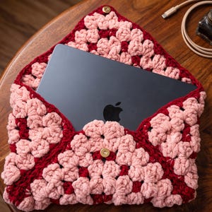 Puede incluir: Un portátil gris oscuro con un logotipo de manzana negro está parcialmente insertado en una funda de ganchillo hecha a mano. La funda presenta un patrón de cuadrados de la abuela en tonos alternos de rojo oscuro y rosa claro, con un broche dorado.