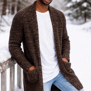 May include: A long-sleeve dark brown knit cardigan with two front pockets, worn over a plain white t-shirt and light blue denim jeans, paired with a light gray ribbed beanie cap. The garment features a thick, textured yarn construction.