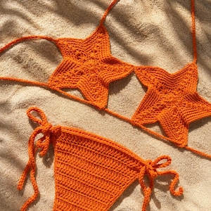 Può includere: Un set bikini all'uncinetto color arancione brillante composto da un top con due coppe a forma di stella collegate da sottili cordini e uno slip abbinato con laccetti laterali. Il capo presenta un motivo a maglia testurizzato ben visibile.