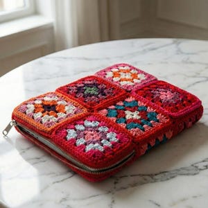Pode incluir: Uma capa de laptop vermelha de crochê com fecho de zíper. A capa é feita de quadrados de vovó coloridos em tons de laranja, rosa, roxo e verde-azulado. A capa está em uma mesa de mármore.
