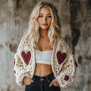 Op de afbeelding: Een crèmekleurige gehaakte cardigan met rode hartachtige accenten. De cardigan heeft een losse pasvorm en lange mouwen.