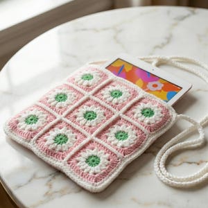 Puede incluir: Una funda de tableta de ganchillo rosa, blanca y verde con un patrón de margarita. La funda tiene una correa blanca y contiene una tableta con una pantalla colorida. La funda está hecha de hilo y tiene forma cuadrada.
