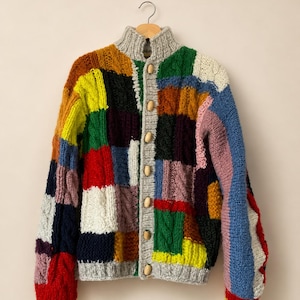 Può includere: Un cardigan patchwork colorato con collo alto e bottoni in legno. Il maglione presenta una varietà di colori, tra cui rosso, blu, verde, giallo e bianco. L'indumento è esposto su una gruccia di legno.