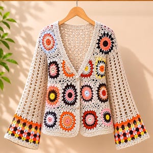 Pode incluir: Um cardigã de crochê bege com decote em V e mangas compridas. A peça é construída a partir de múltiplos motivos quadrados, cada um contendo um padrão floral circular em cores vibrantes, incluindo laranja, rosa, amarelo, preto e azul claro.