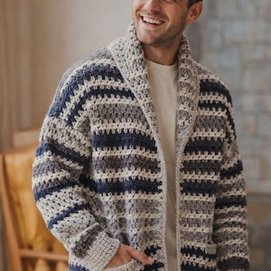 Puede incluir: Cárdigan hecho a mano, con frente abierto, en un patrón de rayas grises, blancas y azul marino. El suéter tiene cuello chal, bolsillos y puños acanalados. El modelo lleva una camiseta blanca debajo.