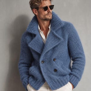 May include: A double-breasted blue knit cardigan with a wide collar, long sleeves, and ribbed cuffs. The garment features a fuzzy, textured surface and two dark buttons on the front, worn over a white collared shirt and light-colored trousers.