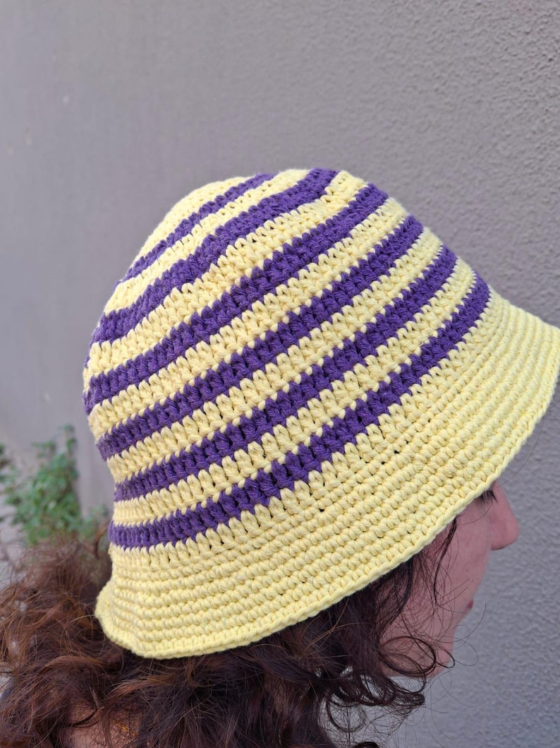 Op de afbeelding: Een handgemaakte gehaakte vissershoed met afwisselende gele en paarse strepen. De hoed is gemaakt van zacht garen en heeft een ronde kroon en een kleine rand. Een stijlvol accessoire voor bescherming tegen de zon.