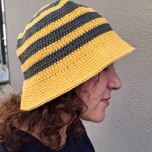 Op de afbeelding: Handgemaakte gehaakte bucket hat met afwisselende horizontale strepen in geel en donkergrijs. De hoed heeft een brede rand en een afgeronde kroon. De hoed is gemaakt van zacht, getextureerd garen.