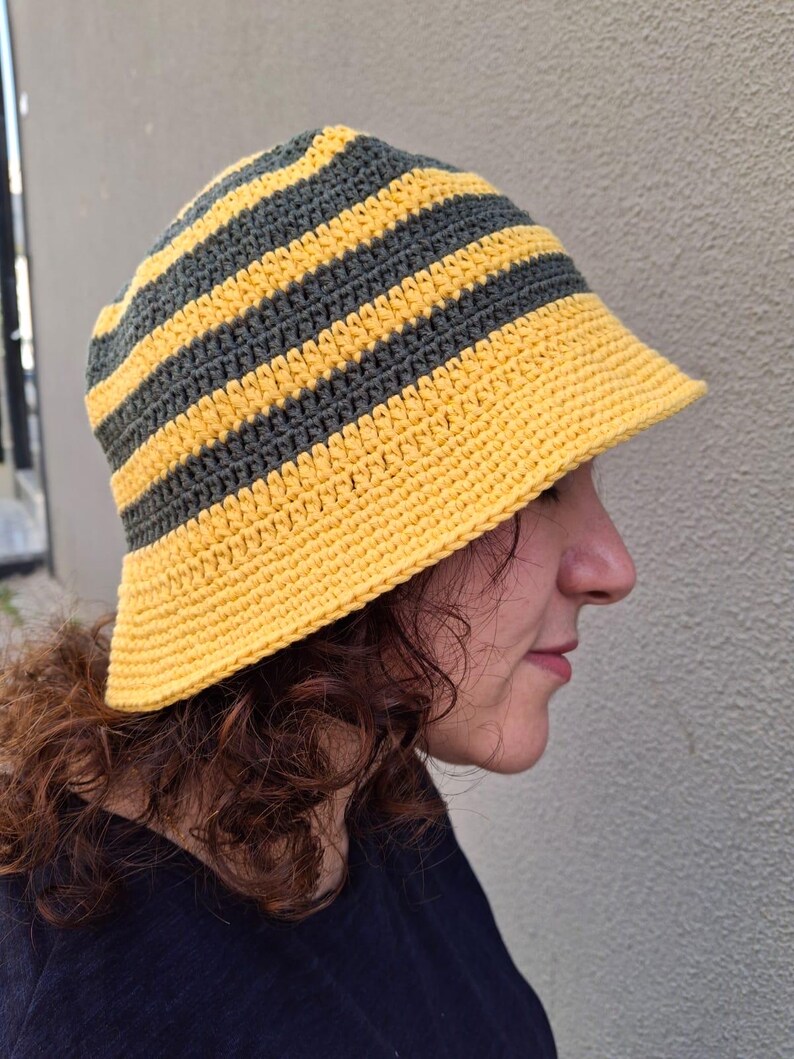 Op de afbeelding: Handgemaakte gehaakte bucket hat met afwisselende horizontale strepen in geel en donkergrijs. De hoed heeft een brede rand en een afgeronde kroon. De hoed is gemaakt van zacht, getextureerd garen.