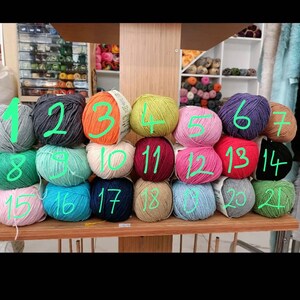 Op de afbeelding: Een display van kleurrijke bollen garen in verschillende tinten, waaronder groen, roze, blauw en oranje. Het garen is gerangschikt op houten planken, met een verscheidenheid aan kleuren en texturen, perfect voor brei- en haakprojecten.