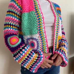 Puede incluir: Una chaqueta de punto de ganchillo colorida con un diseño de patchwork que presenta cuadrados y círculos en tonos de rosa, verde, morado, amarillo y blanco.