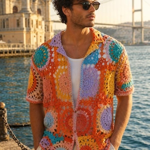 Puede incluir: Una camisa de manga corta con botones hecha de cuadrados de ganchillo multicolores en tonos naranja, amarillo, azul claro, rosa y lavanda. La camisa presenta un cuello clásico, cierre frontal de botones y una textura de punto calado.