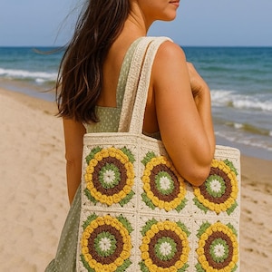 May include: A cream-colored crochet tote bag featuring a sunflower design in yellow, brown, and green. The bag has a rectangular shape with a matching strap. The woman is wearing a green dress and sunglasses, standing on a sandy beach.