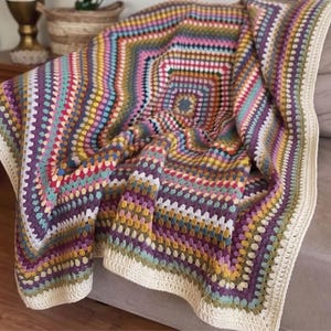 Può includere: Una coperta lavorata all'uncinetto a mano con un vivace motivo geometrico multicolore a quadrati concentrici. Il design presenta sfumature di viola, giallo senape, verde acqua, rosa e blu, rifinita con un bordo color crema a tinta unita.