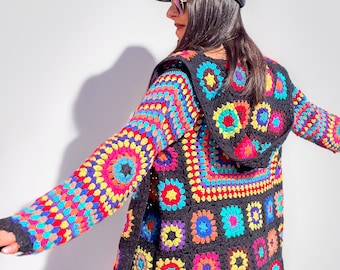 May include: A colorful crochet cardigan with a hood, featuring a black background and multi-colored circular patterns. The cardigan is worn by a person wearing a black cap and black pants.