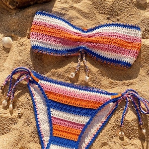 Puede incluir: Un conjunto de bikini de ganchillo de dos piezas con rayas horizontales en tonos vibrantes de azul, blanco, naranja y rosa. La parte superior tipo bandeau y la braga con lazos laterales incluyen cordones trenzados y pequeñas conchas blancas.