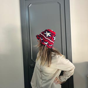 Pode incluir: Um chapéu de balde de crochê vermelho, branco e azul marinho com um padrão geométrico. O chapéu tem uma aba e é feito de fio texturizado. O chapéu é um acessório elegante.