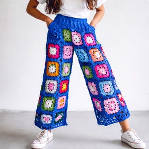 Könnte beinhalten: Blaue Häkelhose mit bunten Quadratmustern. Die Quadrate sind in Rosa, Weiß, Grün und Orange gehalten. Die Hose hat einen hohen Bund und weite Beine. Dazu werden weiße Turnschuhe getragen.