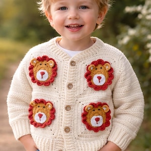 Könnte beinhalten: Cremefarbene Strickjacke mit Knöpfen und vier gehäkelten Löwen-Applikationen. Die Löwen-Applikationen haben eine rote Mähne, ein hellbraunes Gesicht und weiße und rote Details. Die Strickjacke wird von einem Kind mit Jeans getragen.