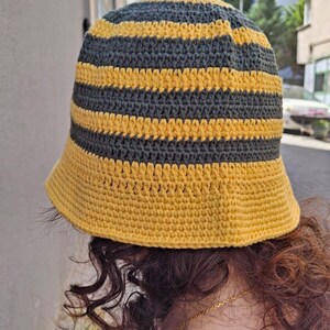 Op de afbeelding: Een handgemaakte gehaakte bucket hat met afwisselende horizontale strepen in geel en donkergrijs. De hoed heeft een brede rand en een ronde kroon. Het garen lijkt een zacht, getextureerd materiaal te zijn. De hoed is ontworpen om gedragen te worden.