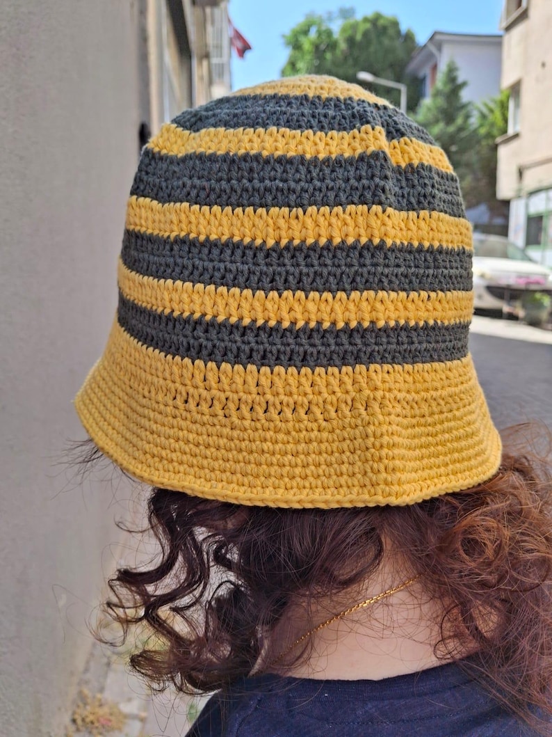 Op de afbeelding: Een handgemaakte gehaakte bucket hat met afwisselende horizontale strepen in geel en donkergrijs. De hoed heeft een brede rand en een ronde kroon. Het garen lijkt een zacht, getextureerd materiaal te zijn. De hoed is ontworpen om gedragen te worden.