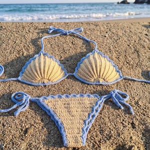 May include: A two-piece crochet bikini set featuring a beige shell-shaped top and matching bottom, both trimmed with light blue scalloped edges. The top has a halter neck tie, and the bottom features side ties for an adjustable fit.