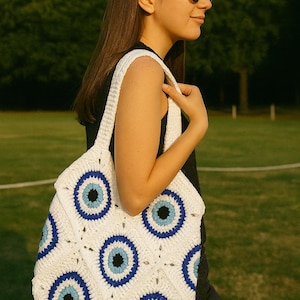 May include: A white, crocheted tote bag featuring a repeating pattern of blue and black eye motifs. The bag, with a square design and shoulder strap, is held by a person. The person is wearing sunglasses and a black top.