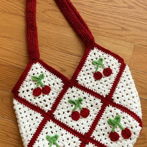 Peut inclure: Sac fourre-tout en crochet fait à la main avec une poignée et une bordure rouges. Le sac présente un fond blanc avec des carrés rouges et des cerises brodées. Les cerises ont des fruits rouges et des tiges vertes. Le sac est sur une surface en bois.
