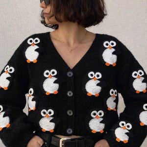 Può includere: Cardigan nero con applicazioni di pinguini bianchi e arancioni. Il maglione con scollo a V ha un'abbottonatura frontale e maniche lunghe. Il maglione è indossato con pantaloni neri e una cintura nera.