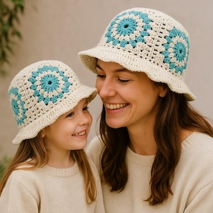 May include: Two cream-colored crochet bucket hats with blue and white floral designs. The hats have a wide brim and are worn by a woman and a child. The hats are handmade and appear to be made of cotton yarn.