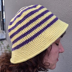 Op de afbeelding: Een gehaakte bucket hat met afwisselende horizontale strepen in geel en paars. De hoed heeft een ronde kroon en een korte rand. De hoed is gemaakt van zacht, getextureerd garen en is een stijlvol accessoire.