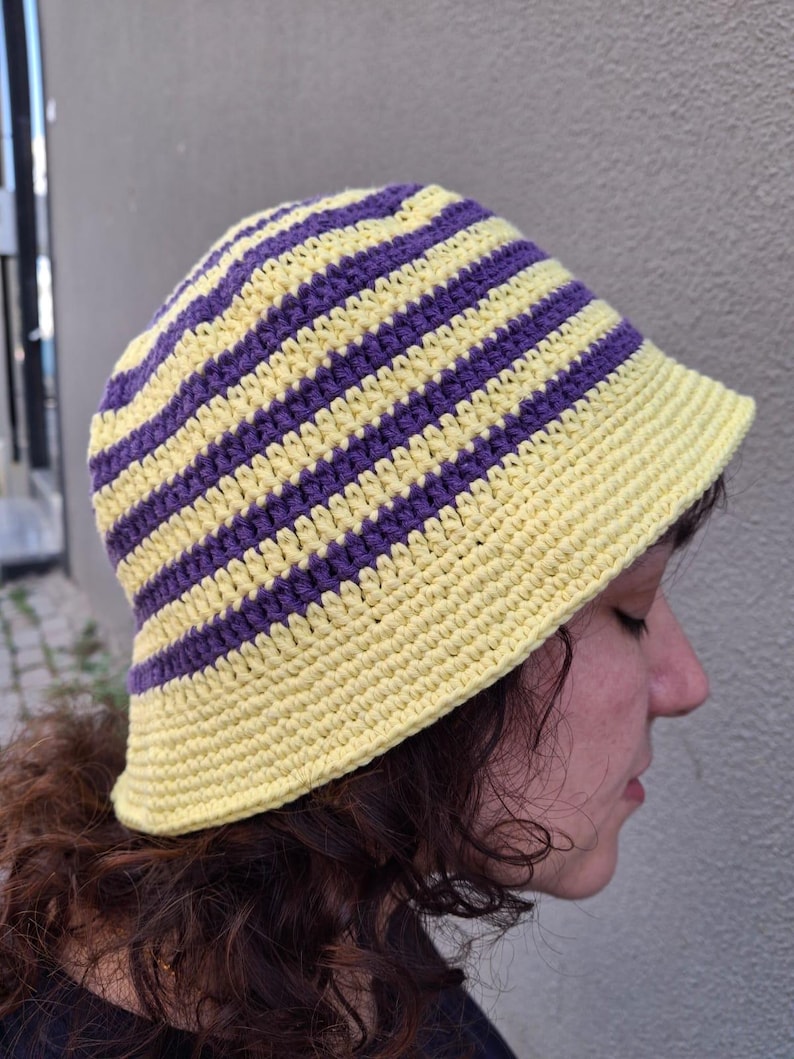 Op de afbeelding: Een gehaakte bucket hat met afwisselende horizontale strepen in geel en paars. De hoed heeft een ronde kroon en een korte rand. De hoed is gemaakt van zacht, getextureerd garen en is een stijlvol accessoire.
