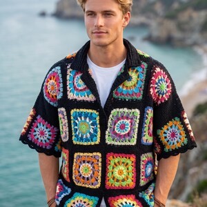 Pode incluir: Uma camisa de manga curta com botões, feita de quadrados de crochê pretos, cada um apresentando um padrão floral multicolorido vibrante em tons de rosa, azul, amarelo, verde e laranja, usada sobre uma camiseta branca lisa.