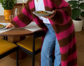 May include: A long, striped cardigan in brown and hot pink is worn over a white t-shirt and blue jeans. The cardigan is the focal point, with its bold color contrast. The person is holding a plate of food, suggesting a dining setting.