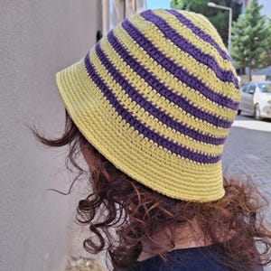 Op de afbeelding: Een handgemaakte gehaakte bucket hat met afwisselende gele en paarse horizontale strepen. De hoed heeft een brede rand en is gemaakt van zacht garen. Deze hoed is een stijlvol accessoire voor bescherming tegen de zon.