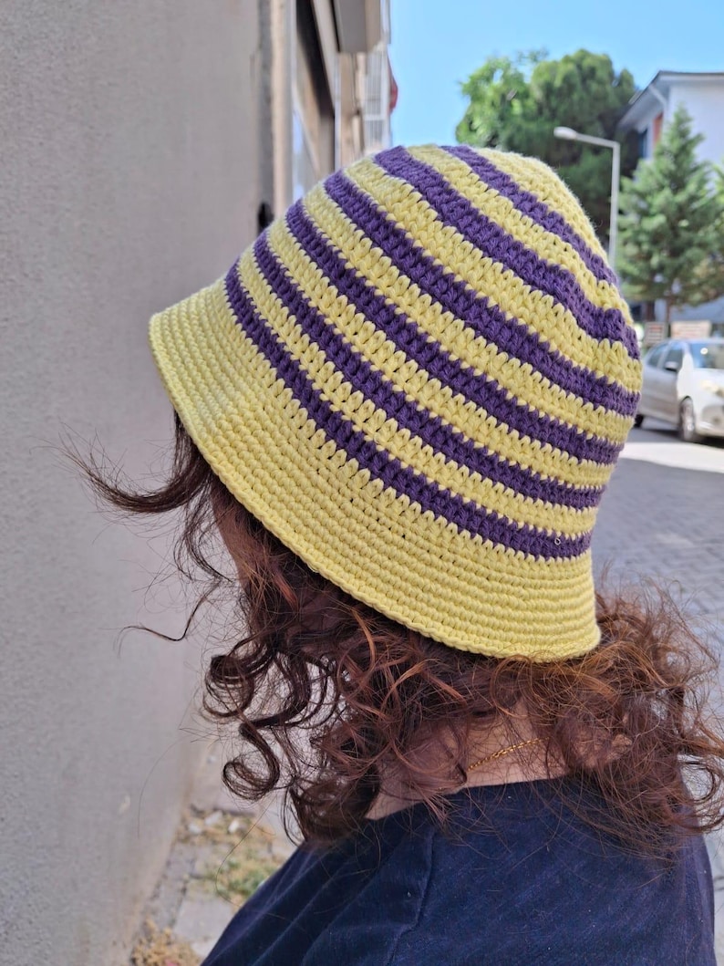 Op de afbeelding: Een handgemaakte gehaakte bucket hat met afwisselende gele en paarse horizontale strepen. De hoed heeft een brede rand en is gemaakt van zacht garen. Deze hoed is een stijlvol accessoire voor bescherming tegen de zon.