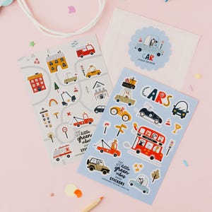 May include: Two rectangular sheets of paper stickers featuring colorful illustrations of cars, buses, and buildings. One sheet has a gray background with a road map design, while the other has a light blue background with the text "CARS" and "little green vibes PAPER STICKERS".