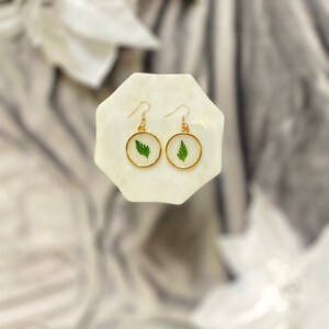 May include: A pair of gold hoop earrings with a small green leaf encased in clear resin. The earrings are displayed on a white marble surface.