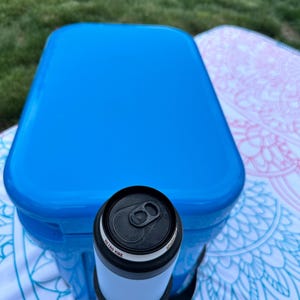 Op de afbeelding: Een blauwe koelbox met een zwart-witte blikjeskoeler. Op de blikjeskoeler staat de tekst "Retro Vibe". De koelbox en de blikjeskoeler liggen op een patroon deken.