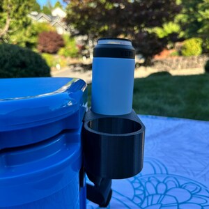 Op de afbeelding: Een zwarte 3D-geprinte bekerhouder bevestigd aan een blauwe koeler. Een lichtblauwe ge&iuml;soleerde blikhouder met een zwarte rand zit in de bekerhouder. De achtergrond toont een groen gazon en bomen.
