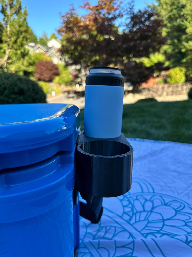 Op de afbeelding: Een zwarte 3D-geprinte bekerhouder bevestigd aan een blauwe koeler. Een lichtblauwe ge&iuml;soleerde blikhouder met een zwarte rand zit in de bekerhouder. De achtergrond toont een groen gazon en bomen.