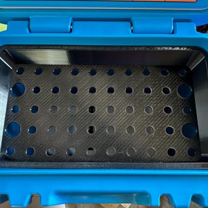 May include: Open, empty blue container with a black interior. The interior features a perforated, dark gray tray. The container is rectangular with rounded corners and a hinged lid.