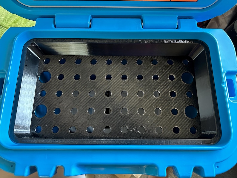 May include: Open, empty blue container with a black interior. The interior features a perforated, dark gray tray. The container is rectangular with rounded corners and a hinged lid.