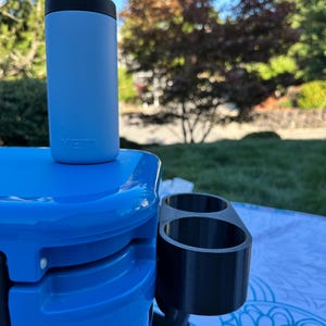 Op de afbeelding: Een lichtblauwe ge&iuml;soleerde blikjeskoeler met een zwarte bovenkant, rustend op een blauwe koeler. Een zwarte bekerhouder is aan de koeler bevestigd. De sc&egrave;ne speelt zich buiten af op een zonnige dag.