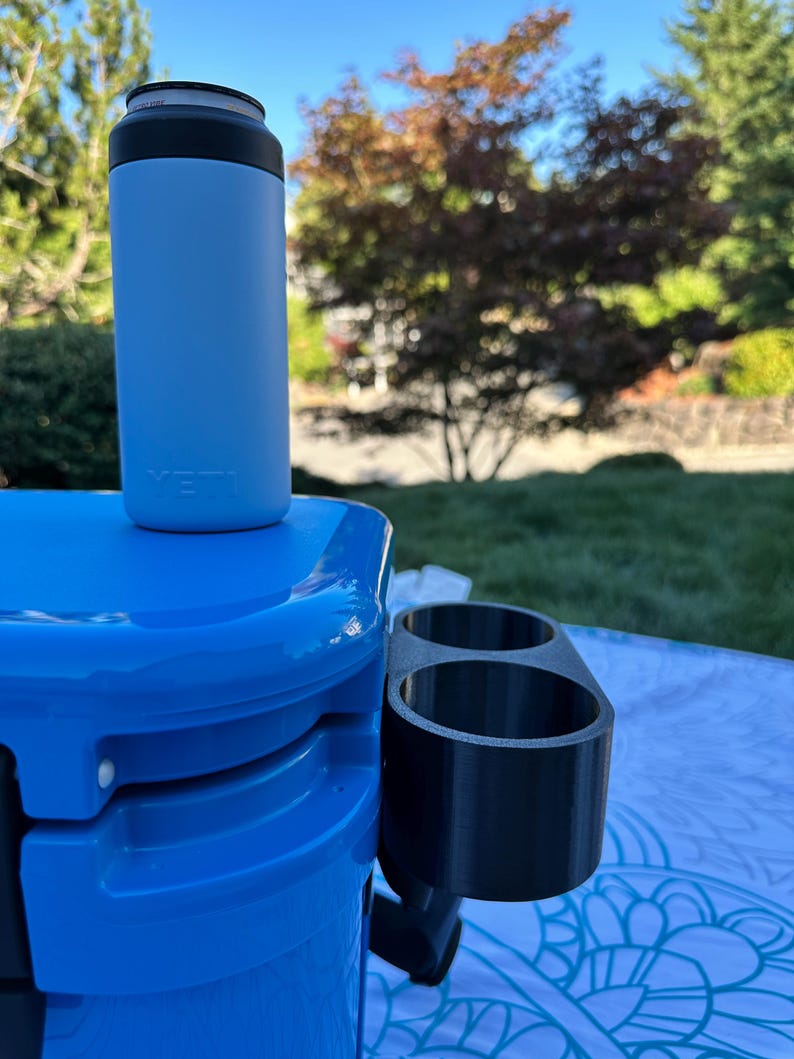 Op de afbeelding: Een lichtblauwe ge&iuml;soleerde blikjeskoeler met een zwarte bovenkant, rustend op een blauwe koeler. Een zwarte bekerhouder is aan de koeler bevestigd. De sc&egrave;ne speelt zich buiten af op een zonnige dag.