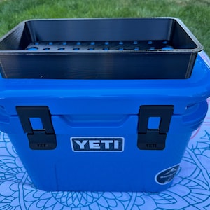 May include: A blue YETI cooler with a black tray on top. The cooler has black latches and the YETI logo on the front. The tray has several holes and is designed to fit the cooler. The cooler is sitting on a patterned surface.