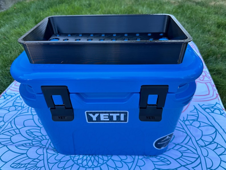 May include: A blue YETI cooler with a black tray on top. The cooler has black latches and the YETI logo on the front. The tray has several holes and is designed to fit the cooler. The cooler is sitting on a patterned surface.