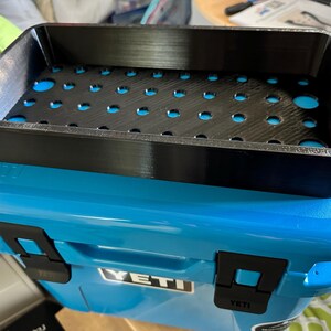 May include: A black, rectangular tray with numerous circular cutouts sits atop a blue cooler. The tray has a textured surface and is designed to fit inside the cooler. The cooler has black latches and the brand name "YETI" is visible.