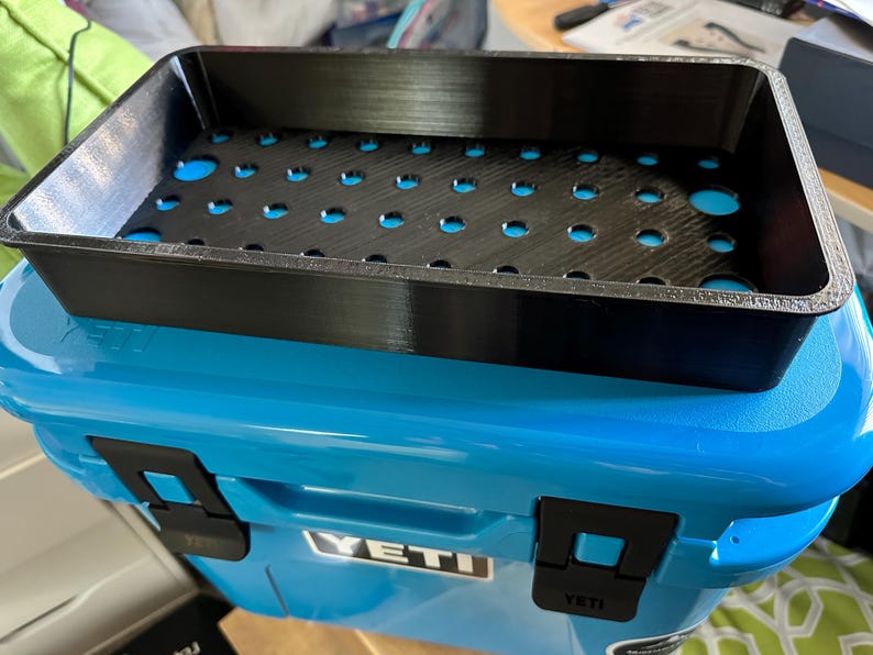 May include: A black, rectangular tray with numerous circular cutouts sits atop a blue cooler. The tray has a textured surface and is designed to fit inside the cooler. The cooler has black latches and the brand name "YETI" is visible.